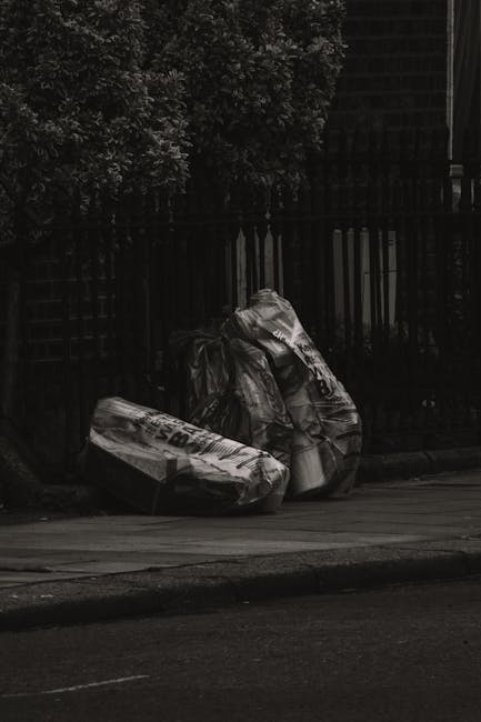 A pile of discarded cardboard boxes and paper waste, including flattened and partially collapsed boxes, resting on the ground next to a rough-textured brick and concrete wall. The cardboard varies in