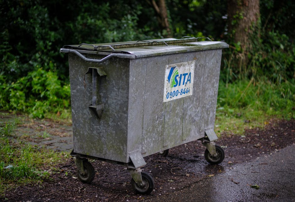 A large, rusty red metal dumpster with vertical ridges on its sides is positioned against the corner of a building, situated on a paved sidewalk. The dumpster's paint is chipped and weathered, showing