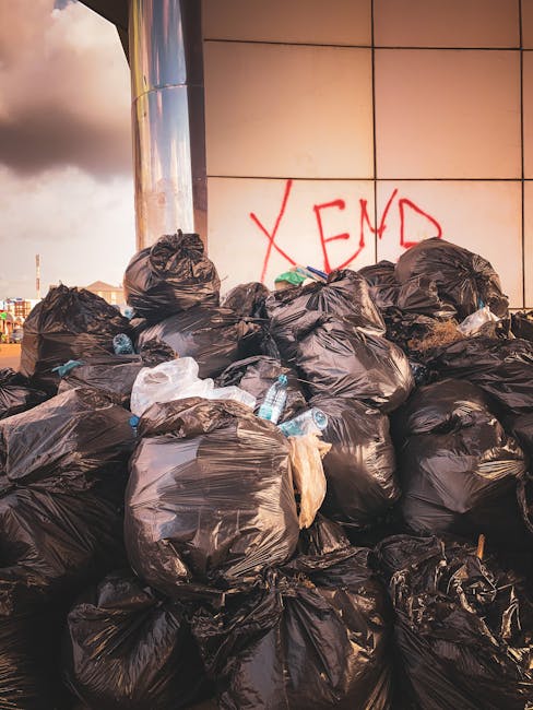 A large pile of black plastic rubbish bags, some tied tightly and others loosely sealed, is stacked against a modern building with a reflective grey and cream tiled facade. The bags appear full and bu